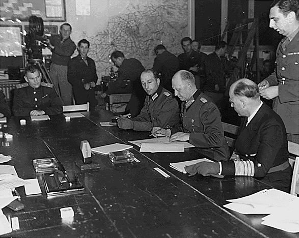 Le Generaloberst Alfred Jodl signant les documents de la capitulation à Reims le 7 mai 1945, pour le Haut Commandement de la Wehrmacht. À sa droite, le Major Wilhelm Oxenius, interprète pour Jodl ; à sa gauche de profil arrière, le Generaladmiral von Friedeburg, commandant en chef de la Kriegsmarine ; derrière lui supervisant les signatures, le Major General Kenneth Strong, du SHAEF ; au fond de face, le colonel Ivan Zenkov, aide de camp, Union soviétique.